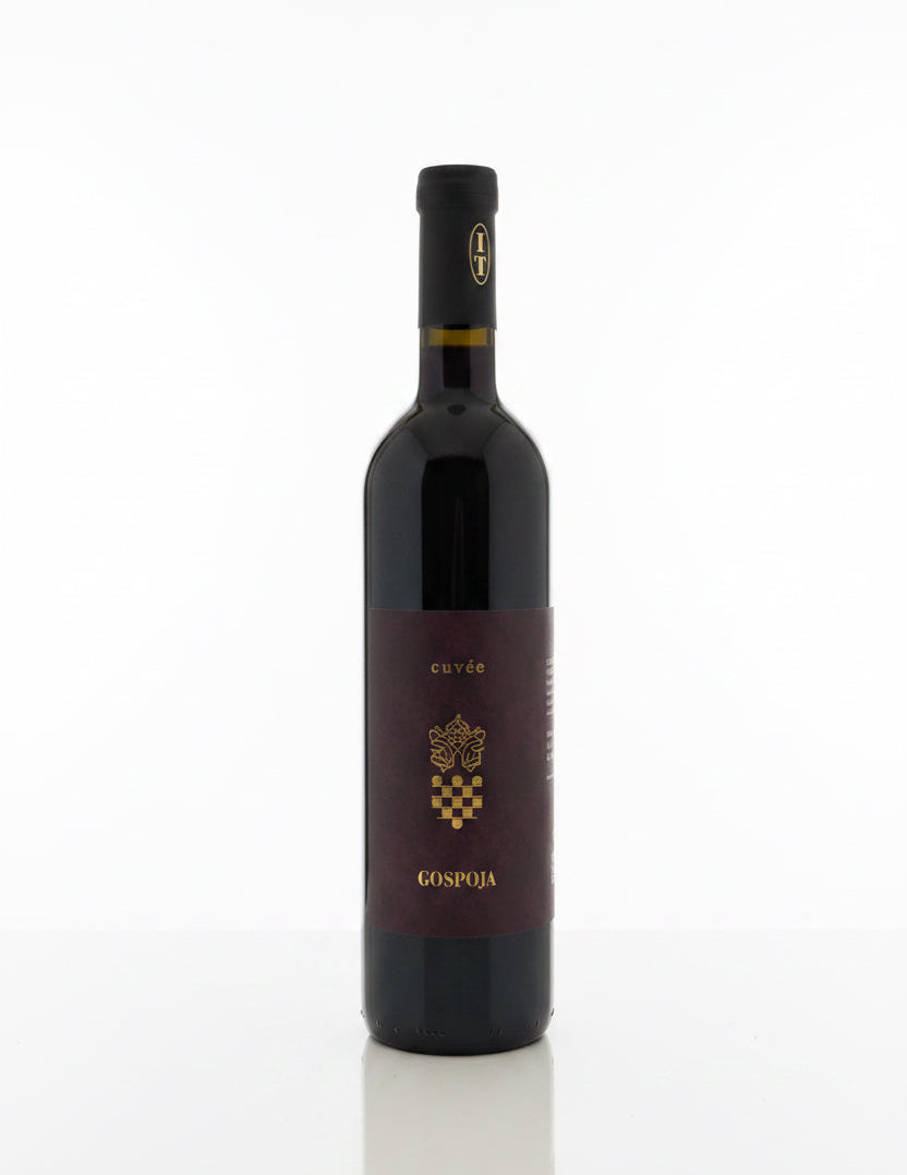 A bottle of Gospoja Cuvés red wine stands on a cardboard box in a warehouse setting, with other bottles and shelving visible in the background.