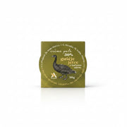 A small square product package with a metallic gold lid featuring a black bird illustration and text, placed on a wooden tabletop next to a white lace cloth.